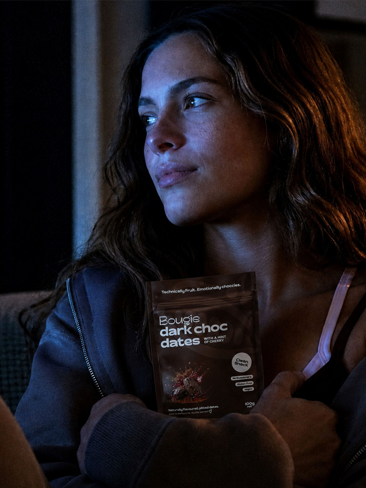 A woman in a dark hoodie holds a pouch of Bougie Dark Chocolate Dates against a dimly lit background.